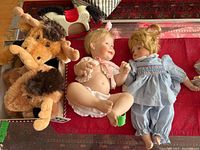Two porcelain dolls, two stuffed moose toys, and one wooden rocking horse toy against a reddish patterned background