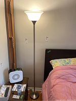 Full view of the tall metal floor lamp beside a bed, showing silver metal stand and white plastic shade.