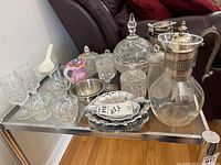 Wide view of the glassware, metal serving dishes, etched candy dish, and glass water pitcher with silverplated details all arranged on a glass table.