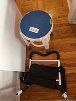 Adjustable shower stool with a blue round seat and four white legs with blue rubber tips, placed near a black and white bed assist bar with a black fabric sling on a wooden floor.