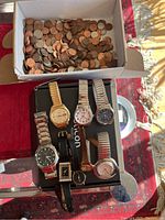 Box of assorted pennies and coins above a collection of 8 watches on a black box, showing varied watch styles and strap types.