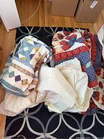 Photo showing pile of folded patchwork quilts including one with red, blue, and cream colors, and white embroidered textiles.
