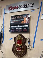 Three advertising items mounted on wall: Coors Light metal sign at top, Shock Top countdown clock sign in middle, Schaefer Falcon wall plaque light at bottom with visible cords.