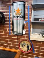 Photo of Sapporo beer advertising light in beer can shape mounted on brick wall, with metal Valley Forge Park tray below on bar ledge.
