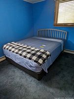 Full view of the bed made with blue fitted sheet, box spring, mattress, and checkered blanket at the foot of the bed, positioned against blue walls and carpet.