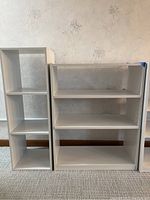 Two white wood-like bookshelves standing side by side against a floral wallpapered wall on a carpeted floor. The bookshelf on the left is taller and narrower with three equal-sized vertical compartments. The bookshelf on the right is wider with three horizontal shelves. Scratches and signs of wear visible on surfaces.