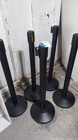 Five black steel pole barriers with weighted bases positioned on concrete floor against a wall.
