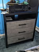 Front view of the dresser showing three drawers with wood look fronts and black handles, items placed on top including electronic equipment.