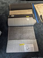 Rolled carpet runners stacked on dark carpet background showing the backing and labels of each; the top one is a woven look rug with a subtle pattern and the bottom one is a plain solid light gray rug.