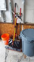 Photo showing lot items arranged in garage: Dirt Devil Vibe Sweeper box, snow shovel, push broom, gray Brute trash can with lid, and orange bucket on concrete floor near plywood wall.