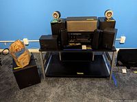 Full view of stereo system with multiple speakers, subwoofer, control center, and bag of wires on a black metal table against a blue wall.