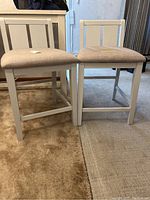 Two bar stools shown side by side on carpet. White painted wood-like frame, beige upholstered seats, backs present.