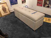 Front angle view of the beige upholstered storage bench showing tufted top and rectangular shape.