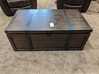 Front view of dark brown wood look storage table with metal bands and stud details across top and sides on carpeted floor.