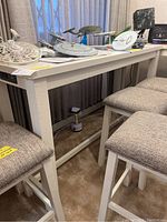 Image of white rectangular bar height table with wood-like finish and upholstered stools around it, items on table not included.