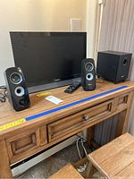 Vizio 22-inch TV screen with two Logitech satellite speakers and one subwoofer speaker on wooden furniture, includes TV remote.