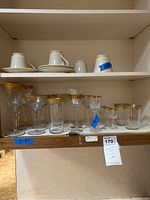 Shelf with 4 white cups, 4 matching saucers, and assorted clear glassware with gold rims.