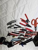Photo showing assorted tools including box cutters, screwdrivers, pliers, scissors and cordless flashlight arranged on white surface.