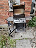 Napoleon gas BBQ grill with three knobs and a side burner with cover, cabinet door open, placed outdoors.
