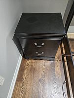 Black-painted veneer night table with two closed drawers against a wall on hardwood floor, showing minimal surface wear.