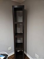 Front view of tall, narrow shelving unit showing four open compartments and dark brown finish, standing upright against a wall.