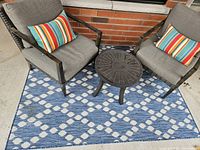 Full patio set including two cushioned chairs with striped pillows, small round metal table, and blue and white patterned carpet on concrete patio next to brick wall.