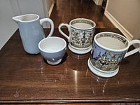Two Emma Bridgewater mugs showing intricate English and heraldic designs and two white ceramic vessels (milk pitcher and sugar bowl) by Marilu on a wooden surface