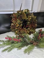 Autumn wreath showing pinecones, oak leaves, red berries, and small yellow flowers, displayed on a bed.