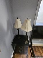 Two brass finished metal lamps with cream colored shades placed on a black surface next to a chair and grey walls.