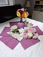 Overview of the table decoration arrangement showing purple glass vase with ranunculus flowers, artificial peonies, candles, ceramic vase, and fabric placemats on a white bedspread.