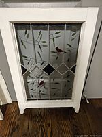 Antique style stained glass art panel with red cardinals and green leaf design in a white wooden frame placed on wooden floor against a wall.