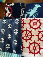 Four assorted nautical themed beach towels folded and laid out showing anchor, lobster, rope, and ship wheel patterns.