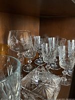 Photo showing assorted crystal stemware and a square lidded dish with cut patterns on a wooden shelf.
