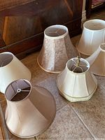 Five assorted lamp shades arranged on floor, varying in size and fabric covering, colors beige, cream, light brown