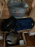 Image showing galvanized bucket on floor and assorted bags including a blue duffel and black JVC bag, alongside a used metal welding mask.