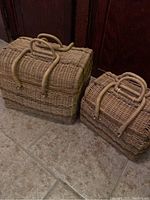 Photo showing all three wicker baskets nested next to each other on tile floor in front of wooden surface. Baskets are rectangular with rounded corners and have tightly woven wicker. Each basket has lid and two handles visible.