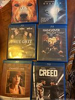 Six Blu-Ray movies including A Dog's Purpose, Gravity, True Grit, Hangover Trilogy, Changeling, and Creed, arranged on a wooden surface.
