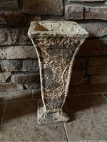Single concrete flower pot or vase stand with rough textured finish shown on tiled floor against stone wall background