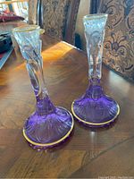 Pair of purple crystal candle holders shown on a wooden table with visible gold trim and decorative design.