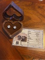 Pair of round clear Cubic Zirconia stone stud earrings in sterling silver settings displayed inside a heart-shaped wooden box alongside a silver quality certificate.