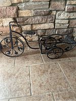 Full view of black metal bicycle-shaped plant stand with decorative scrollwork and basket compartment on tiled floor against stone wall.