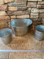 Three metal containers on tile floor against a stone wall background, showing one large hammered metal bin in the center and two smaller galvanized buckets on either side with copper tone handles.