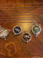 Four dragonfly pendant necklaces with blue, green, and white felt inserts on silver ball chains laid out on wooden surface