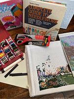 Wide view of needlework lot including books, floss, scissors, craft glue, knitting kit, and unfinished cross-stitch canvas.