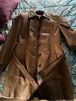 Photo showing full front view of brown leather coat with belt laid flat on surface, displaying collar, button closure, and pocket details.