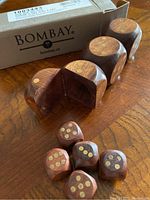 Six wooden dice placed in front of a rectangular wooden holder and the original Bombay box on a wooden surface.