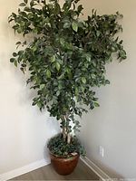 Full view of tall artificial potted tree with broad green leaves in decorative pot placed in corner against walls and floor.