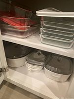 Clear glass Pyrex bakeware pieces stacked and arranged inside a cupboard shelf under a white casserole dish and Corning Ware pans.