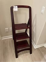 Full view of the dark wood step ladder open and standing upright against a wall on wood floor.