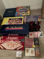 Photo showing assortment of board games including Scrabble, Pictionary, Cranium Deluxe and Booster Box, Chess/Checkers Cabinet, multiple decks of cards and dice cups arranged on floor.